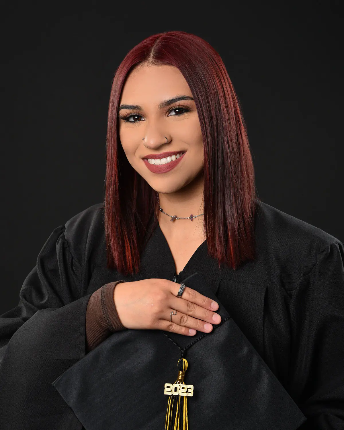 Young woman with straight burgundy-red hair in black graduation gown, smiling, holding her black cap with gold '2023' ta