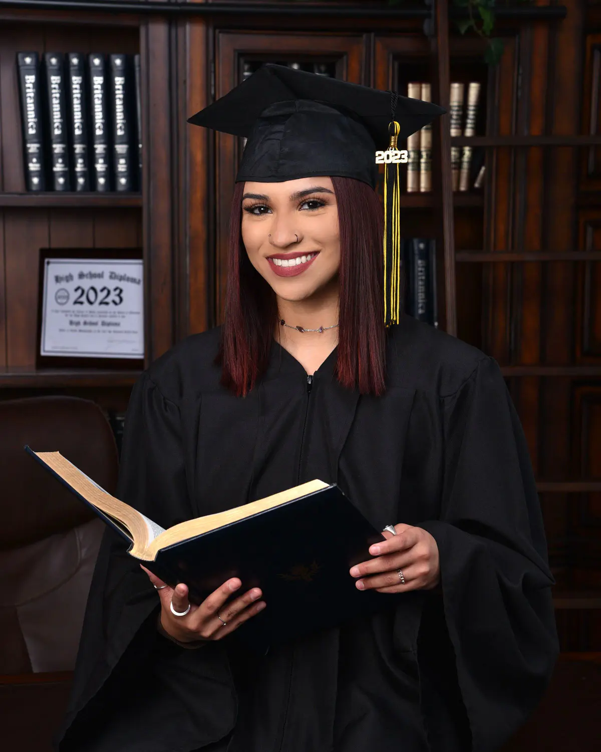 Young woman in black cap and gown with 2023 tassel, reading an open book, seated in library set with dark wood shelves
