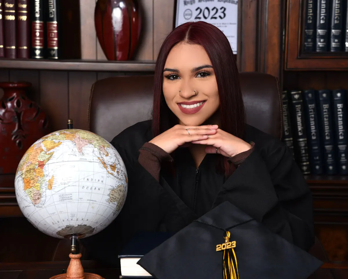 Young woman in black gown at a desk, chin resting on interlaced hands, vintage globe at left, black cap with 2023 tassel on desk, leather chair
