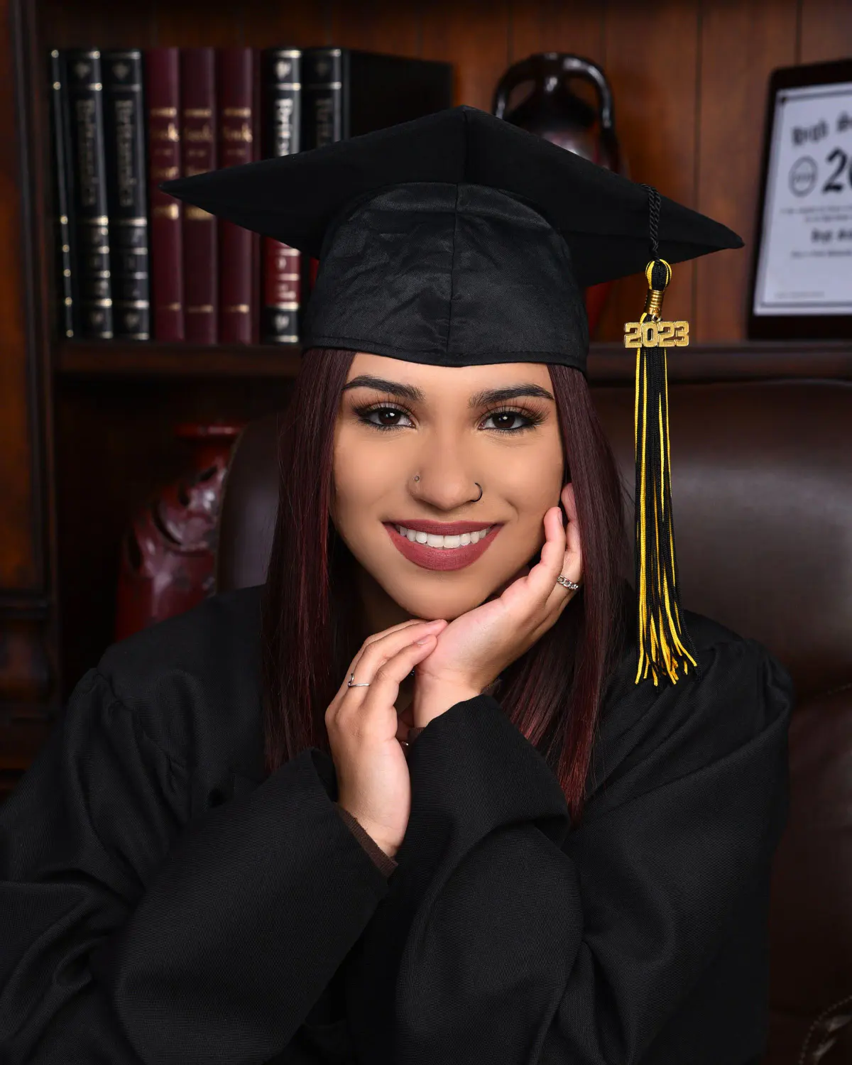 Young woman in black cap with gold 2023 tassel and gown, hands framing her face, big smile, library set with bookshelves