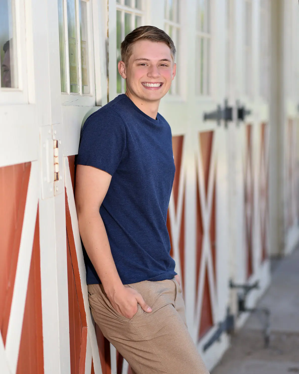 Smiling young man in navy t-shirt and khaki pants leaning against white barn with red-and-white doors and windows, hand in pocket, bright daylight