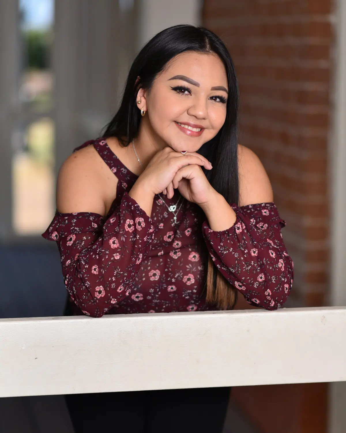 Young woman with long dark hair in burgundy floral off-shoulder top, chin resting on hands on white railing, smiling