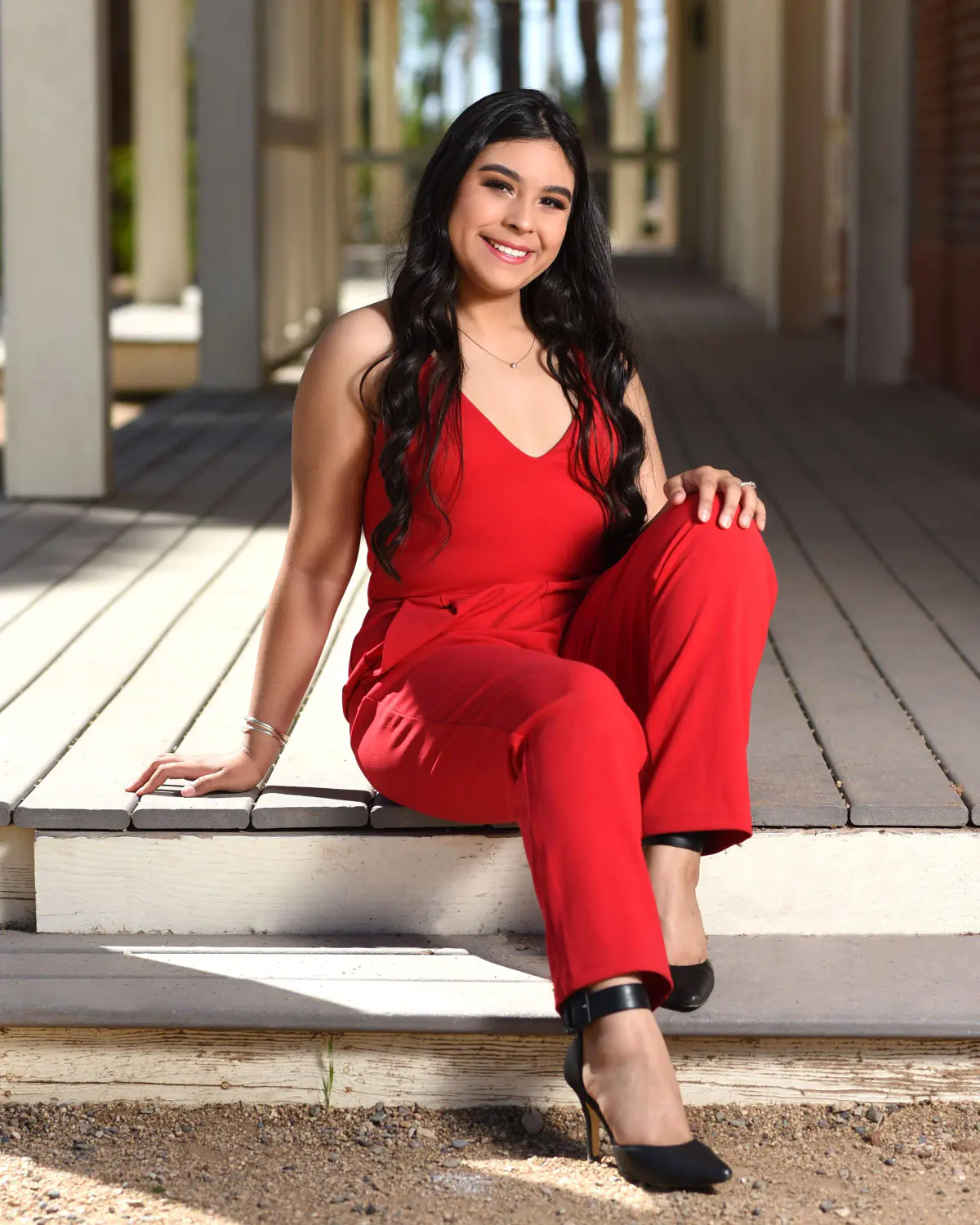 Young woman with long dark hair in a red jumpsuit and black heels, sitting on the edge of a wooden boardwalk/porch with white posts