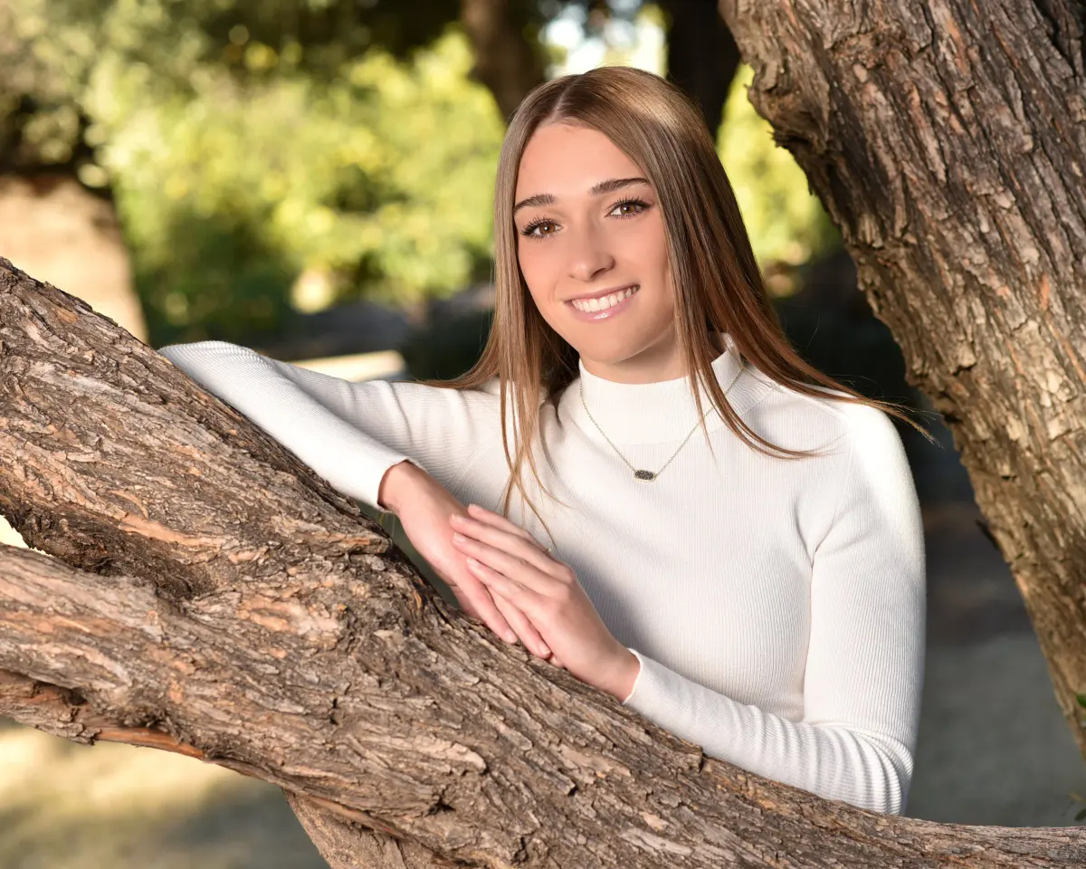 Smiling young woman in a white ribbed mock-neck sweater and fine necklace, leaning on a thick gnarled tree branch, soft green bokeh park background
