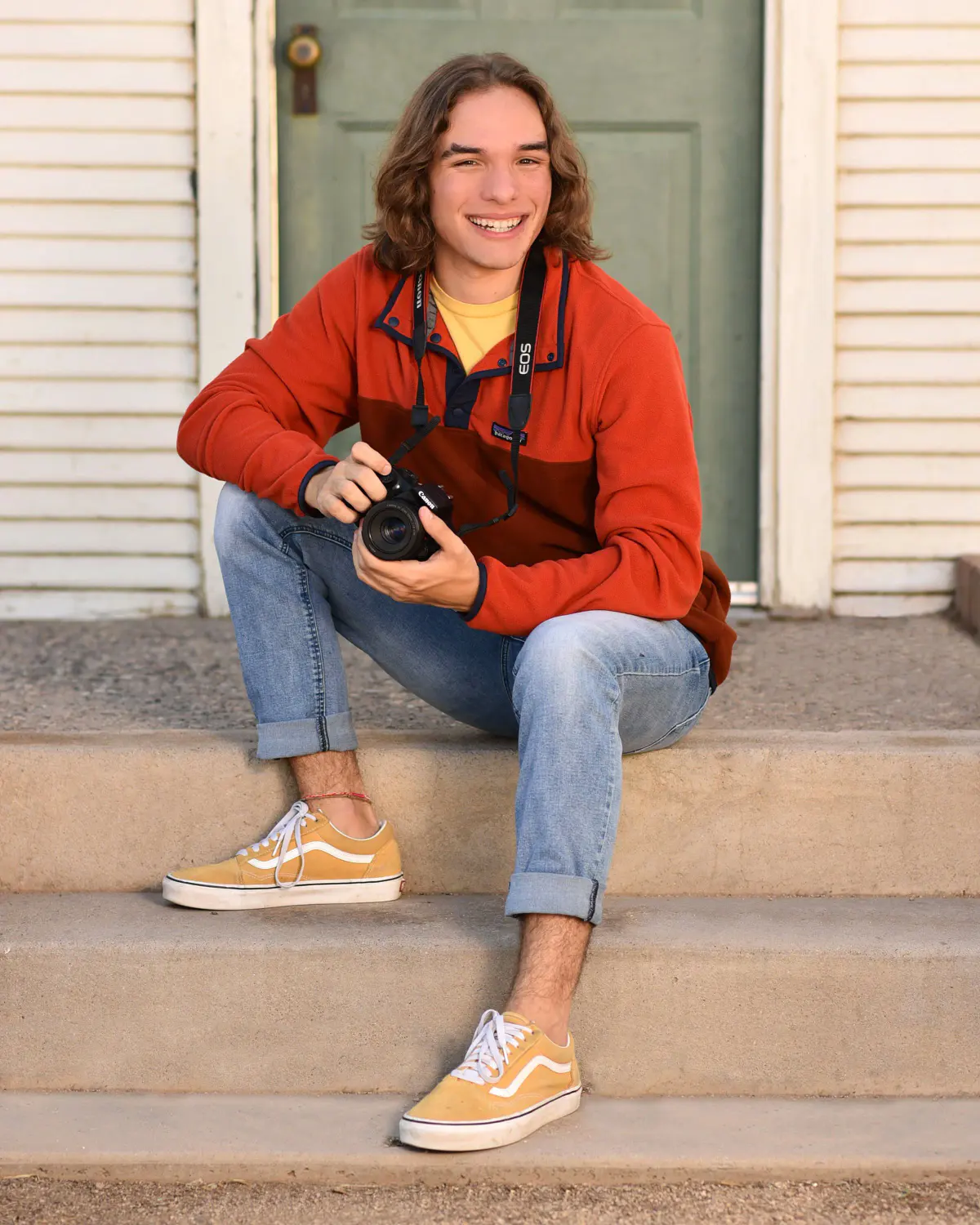 Smiling young man with shoulder-length hair in an orange/red fleece over a yellow tee, jeans and mustard sneakers, holding a DSLR camera
