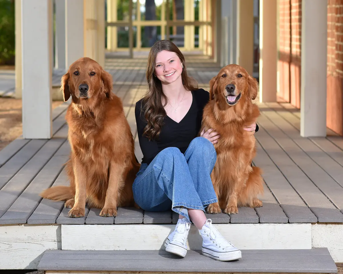 Young woman in a black top, jeans and white sneakers sitting on a wooden porch between two golden retrievers, colonnade receding behind