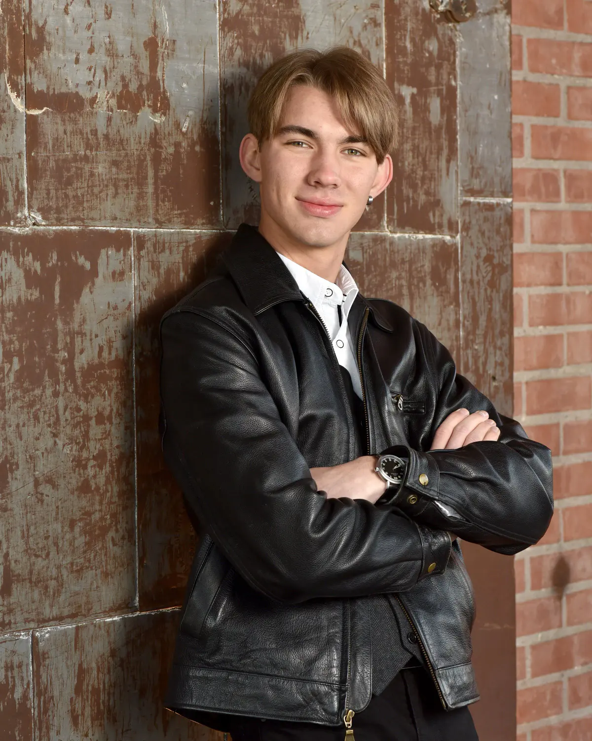 Young man in a black leather jacket over a white shirt, arms crossed, wristwatch, slight smile