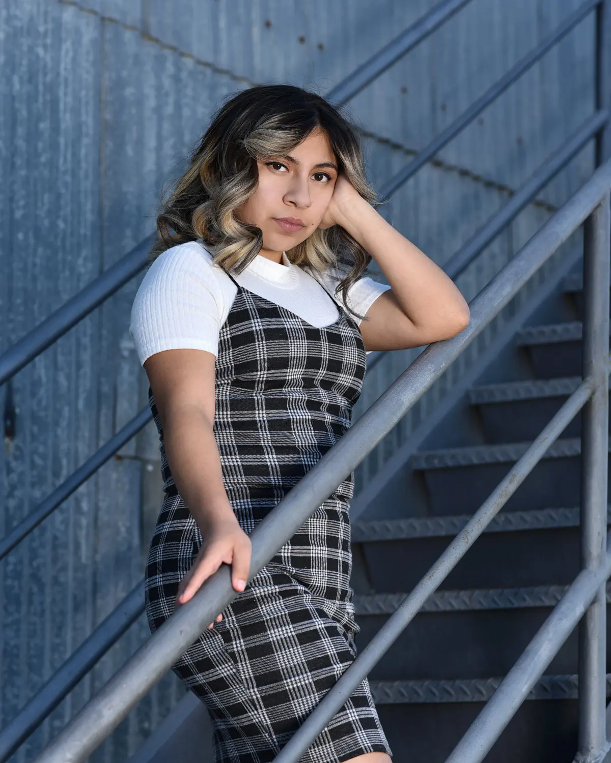 Young woman with highlighted wavy hair, white tee under a black plaid pinafore dress, hand to head