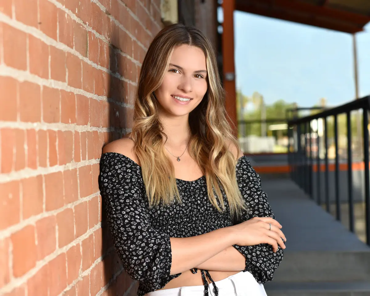 Smiling young woman with long highlighted waves in a black floral off-shoulder top, arms crossed, leaning on a red brick wall