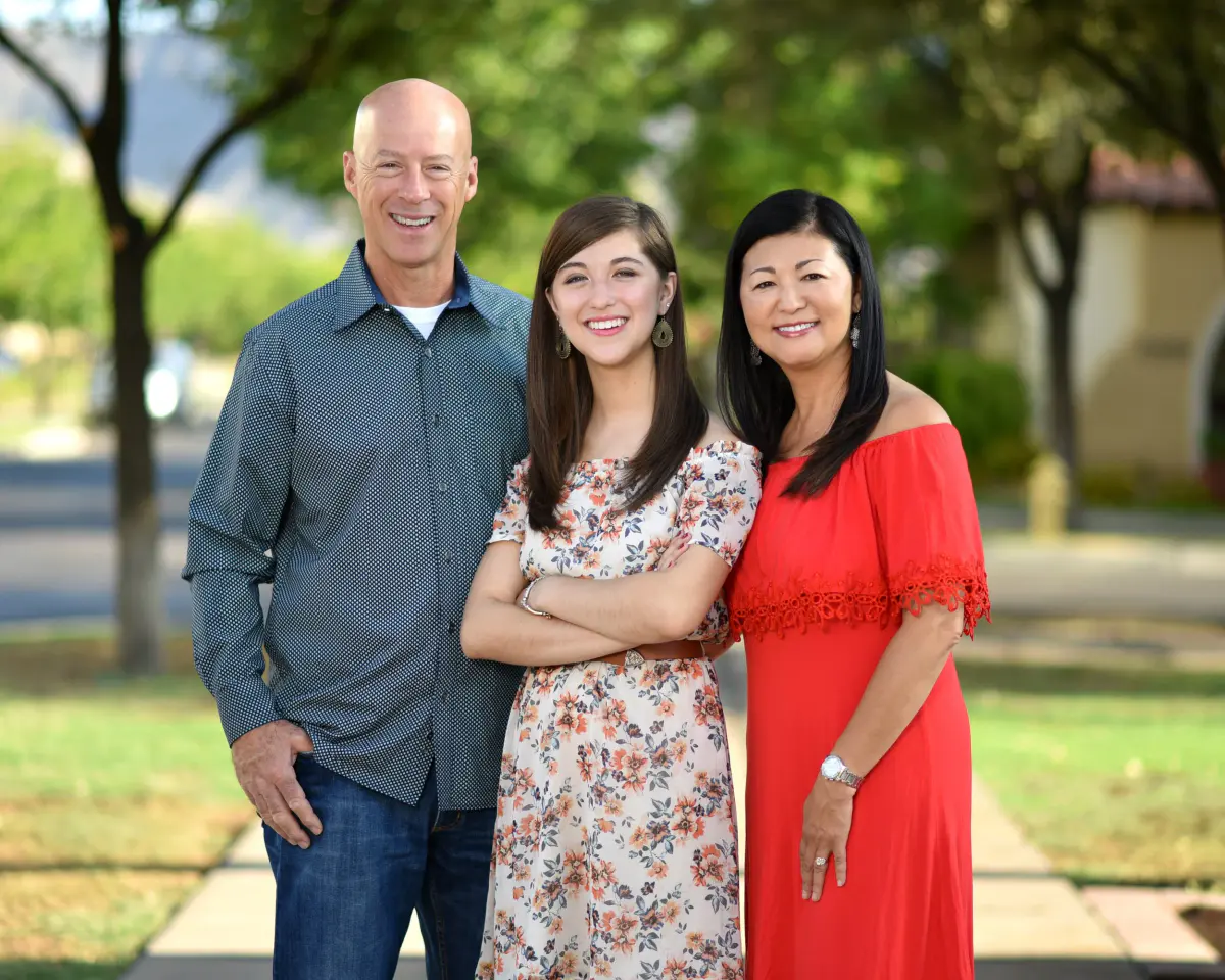 Outdoor family portrait of three adults (an older man, a young woman in a floral dress and an older woman in a red off-shoulder dress) standing on a s