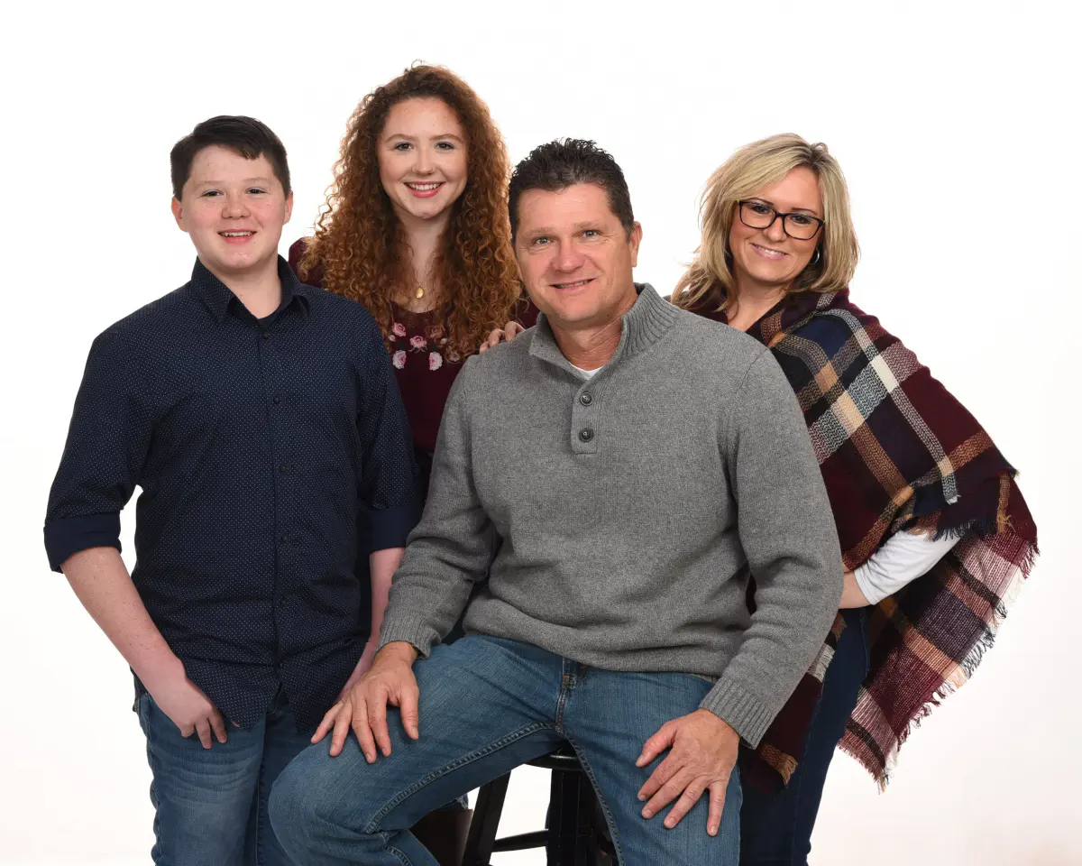 Family of four (teen boy, young woman, seated man in a gray sweater, woman in a plaid wrap) posed on a white seamless background