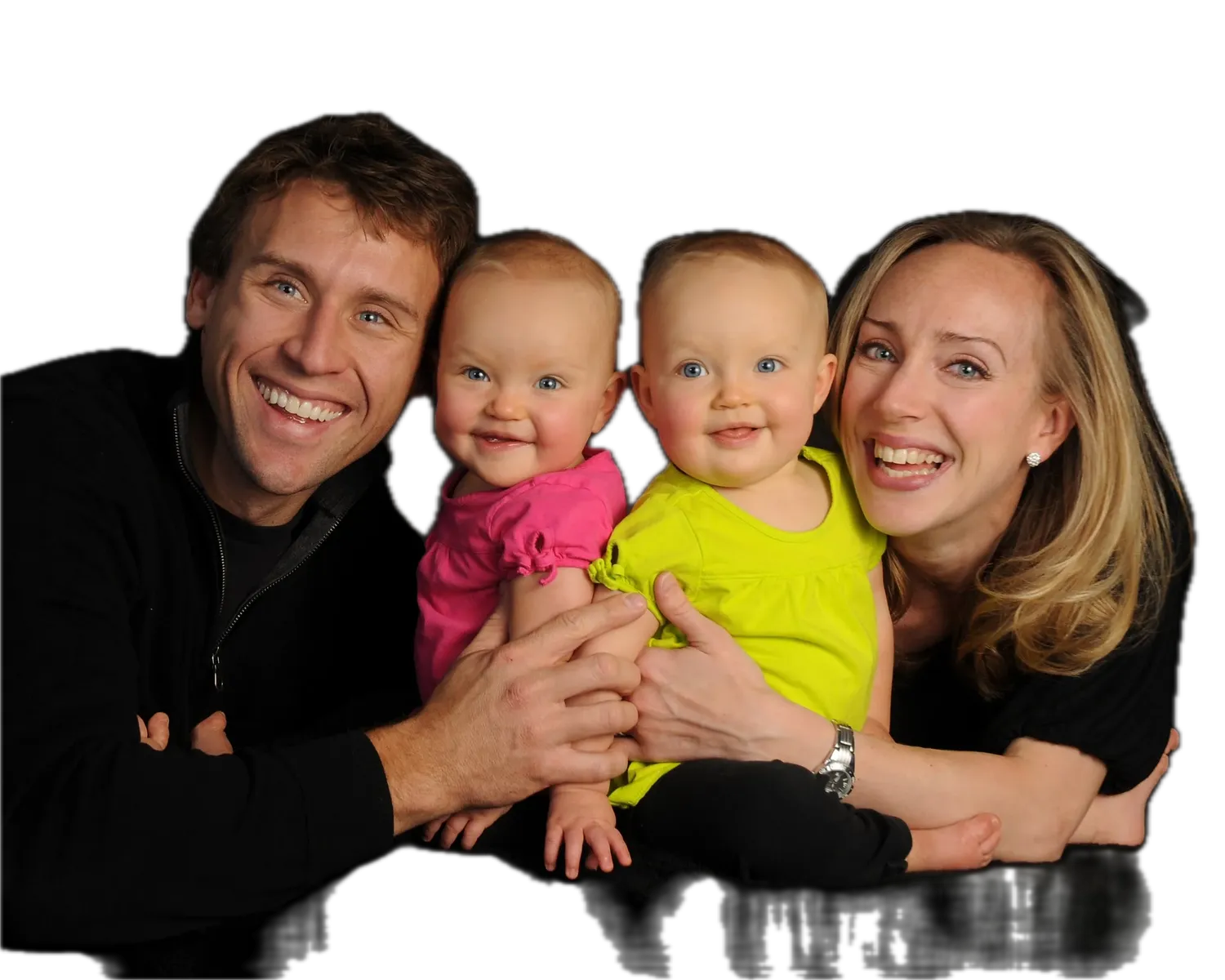 Father and mother on a black backdrop, both smiling broadly while holding twin babies in pink and neon yellow outfits between them