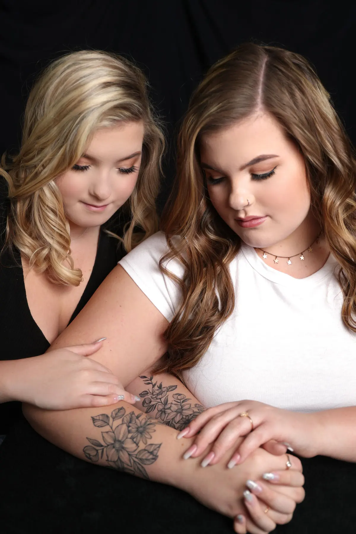 Two young women on a black backdrop, one leaning over the other's shoulder, both looking down at their joined hands
