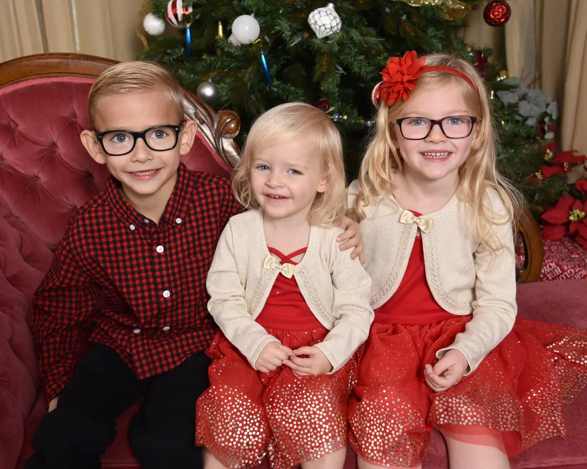 Three young children (a boy in glasses and red plaid, a toddler girl and an older girl in glasses