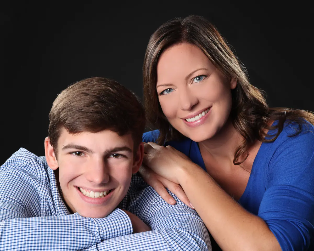 Teen boy in a blue gingham shirt and a woman in a blue top, likely mother and son (not confirmed), posed cheek-to-cheek, smiling, on a black backdrop