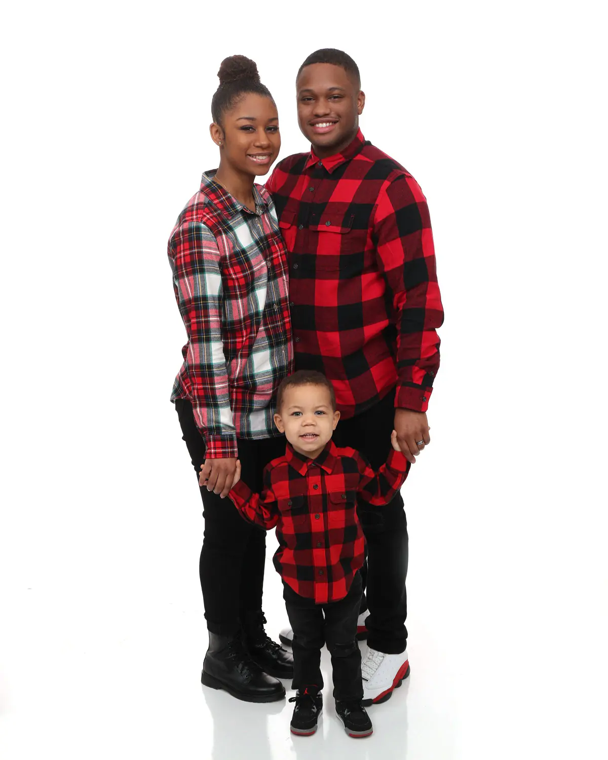 Young couple and a toddler, all in matching red-and-black buffalo plaid shirts, standing full length on white seamless