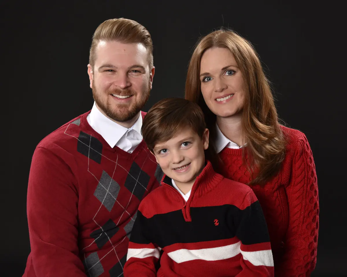 Man, woman and young boy in coordinated red sweaters (argyle, cable knit, striped) posed close together and smiling on a black backdrop