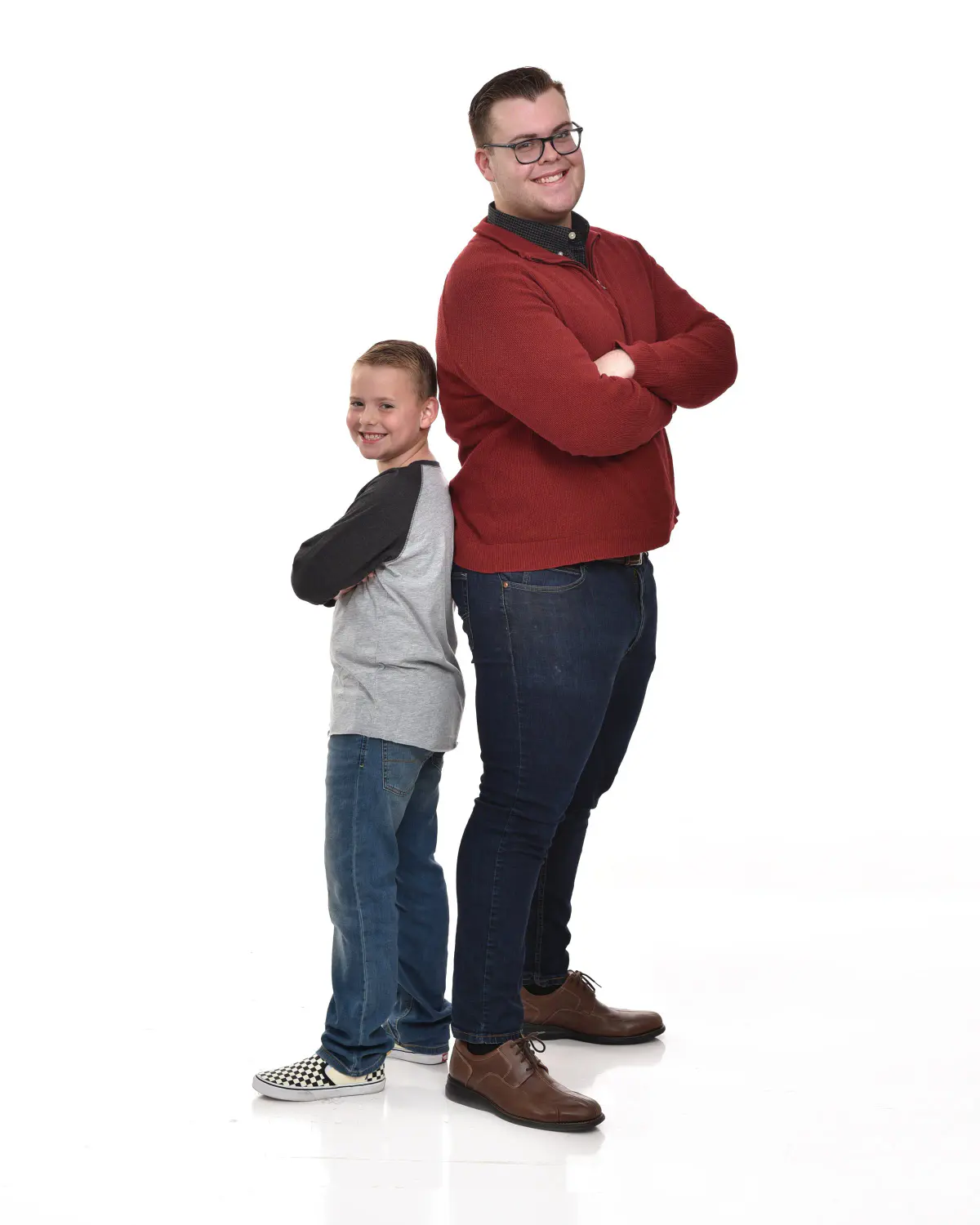 Adult man in a red sweater and a young boy in a raglan tee standing full length with arms crossed, both looking at the camera, on white seamless