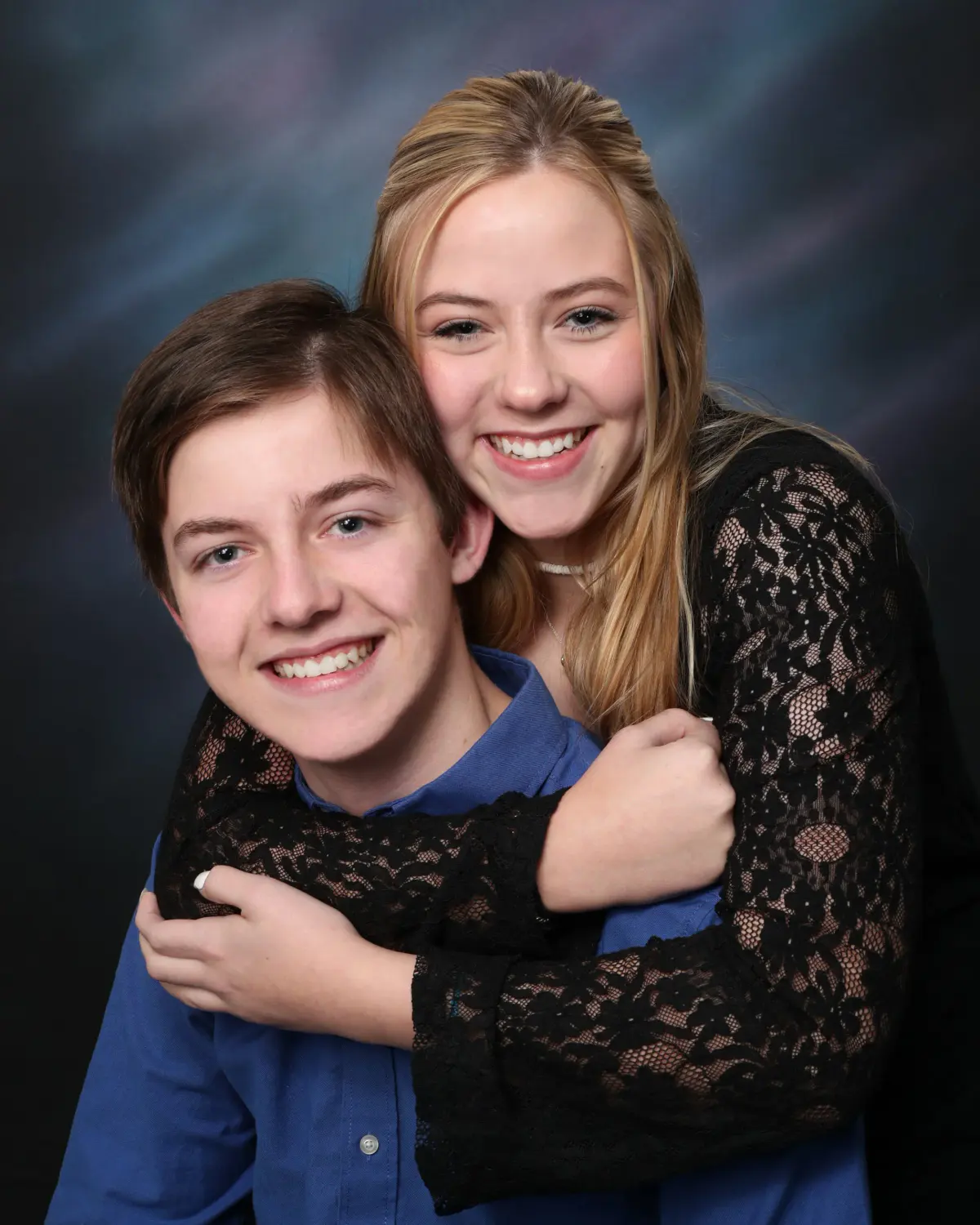 Teen boy and a teen/young-adult woman hugging and smiling, in front of a mottled blue/purple painted canvas backdrop