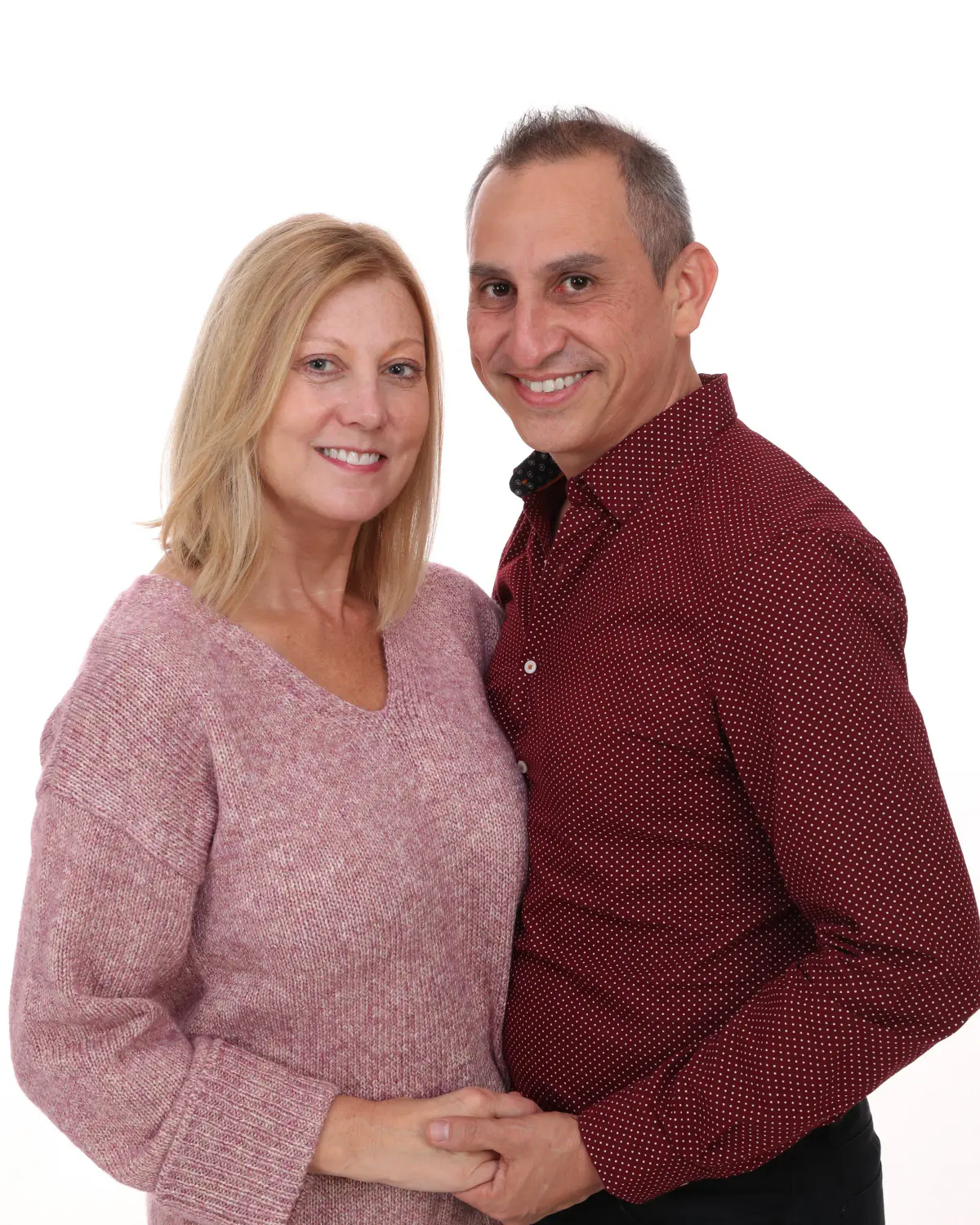 Middle-aged couple embracing, woman in pink sweater, man in burgundy shirt, hands clasped, smiling, on white seamless