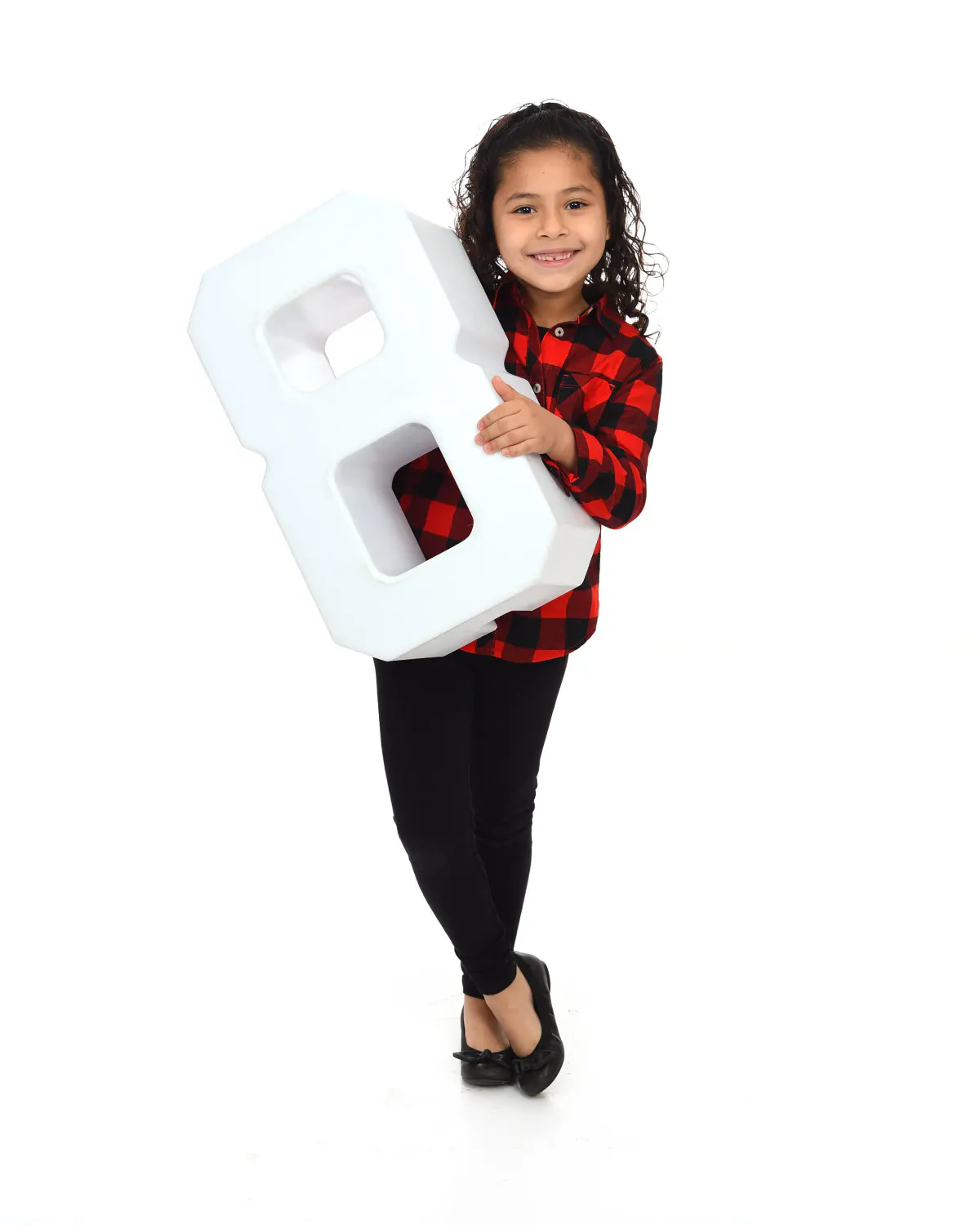 Young girl with curly hair in red plaid shirt and black leggings holding a large white foam numeral '8' prop, standing on white seamless