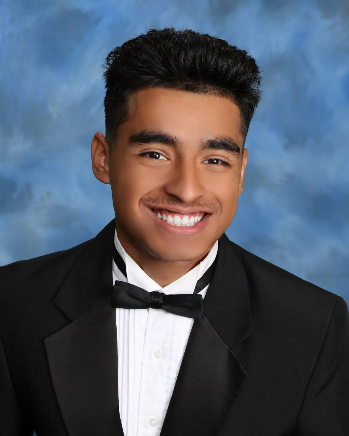 Senior young man, close headshot, big smile, in a black tuxedo with bow tie and pleated white shirt; blue-and-white mottled cloud backdrop