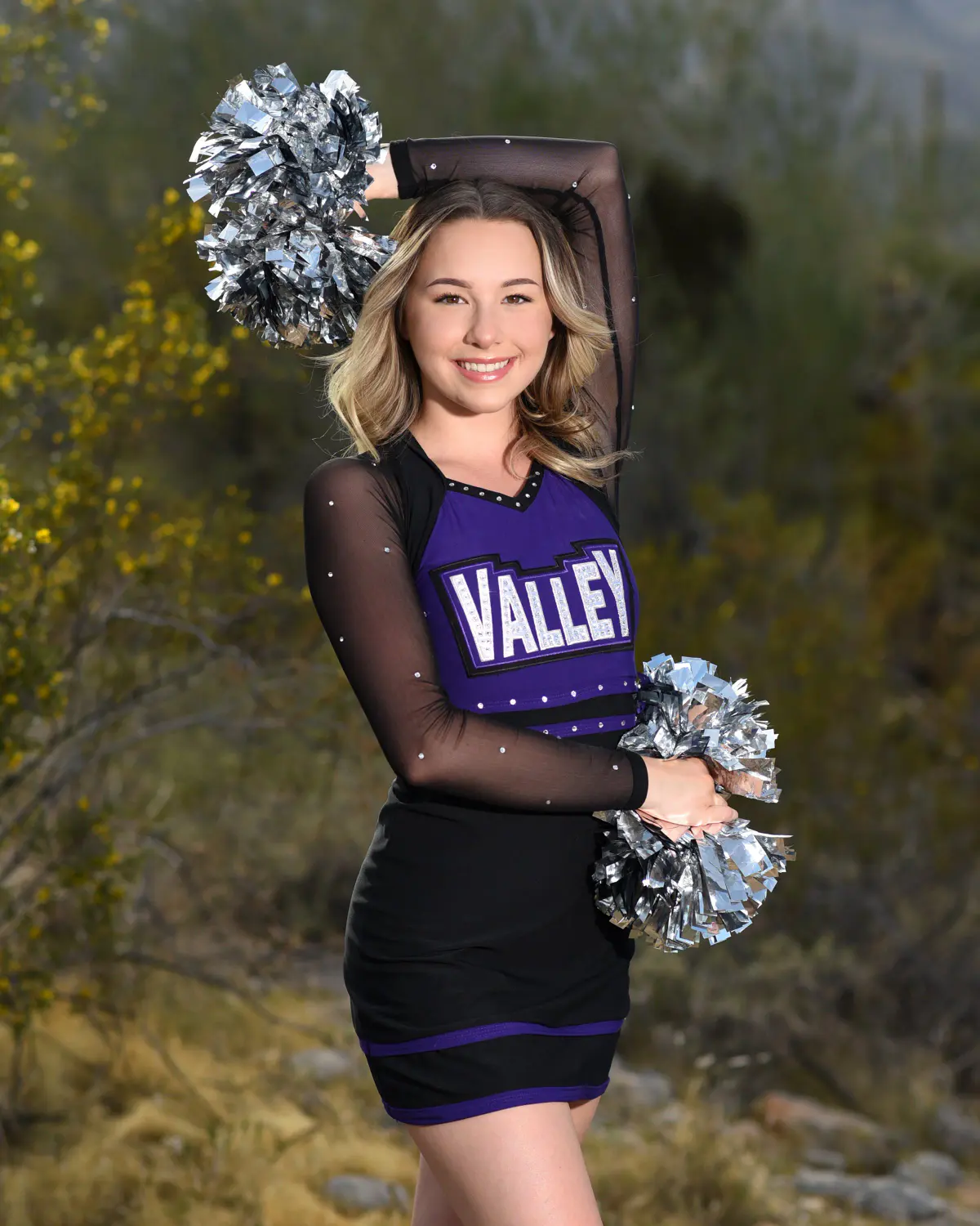 Teen cheerleader in a purple, black and rhinestone 'VALLEY' uniform with sheer sleeves, one arm raised holding a silver pom overhead, other pom at hip