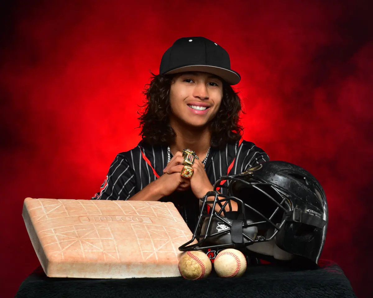 Smiling baseball player in pinstripe jersey and black cap, holding a small item (ring/trophy) at chest, seated behind a black table with a base