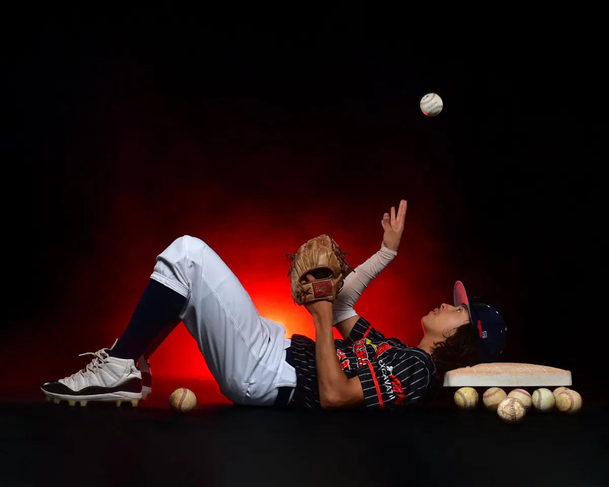 Baseball player lying on his back on a black floor, head on a base, tossing a baseball straight up with glove raised
