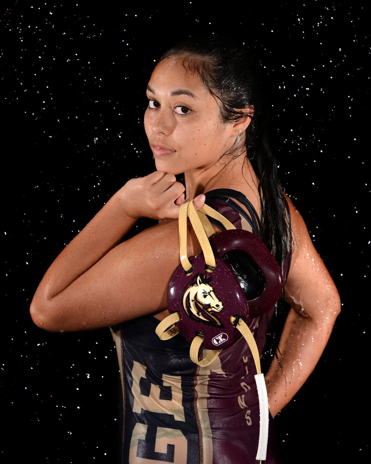 Wrestler in maroon and gold singlet looking back over her shoulder at the camera, holding wrestling headgear with a horse-head logo over her shoulder