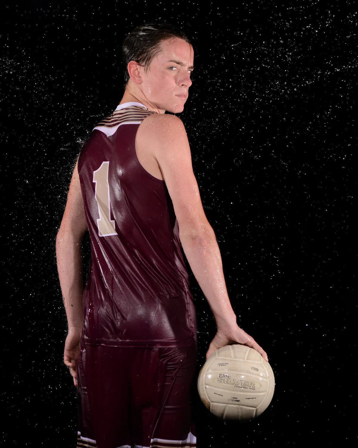 Athlete in wet maroon sleeveless #1 jersey and shorts, back to camera looking over shoulder, holding a cream volleyball at hip, water spray on black