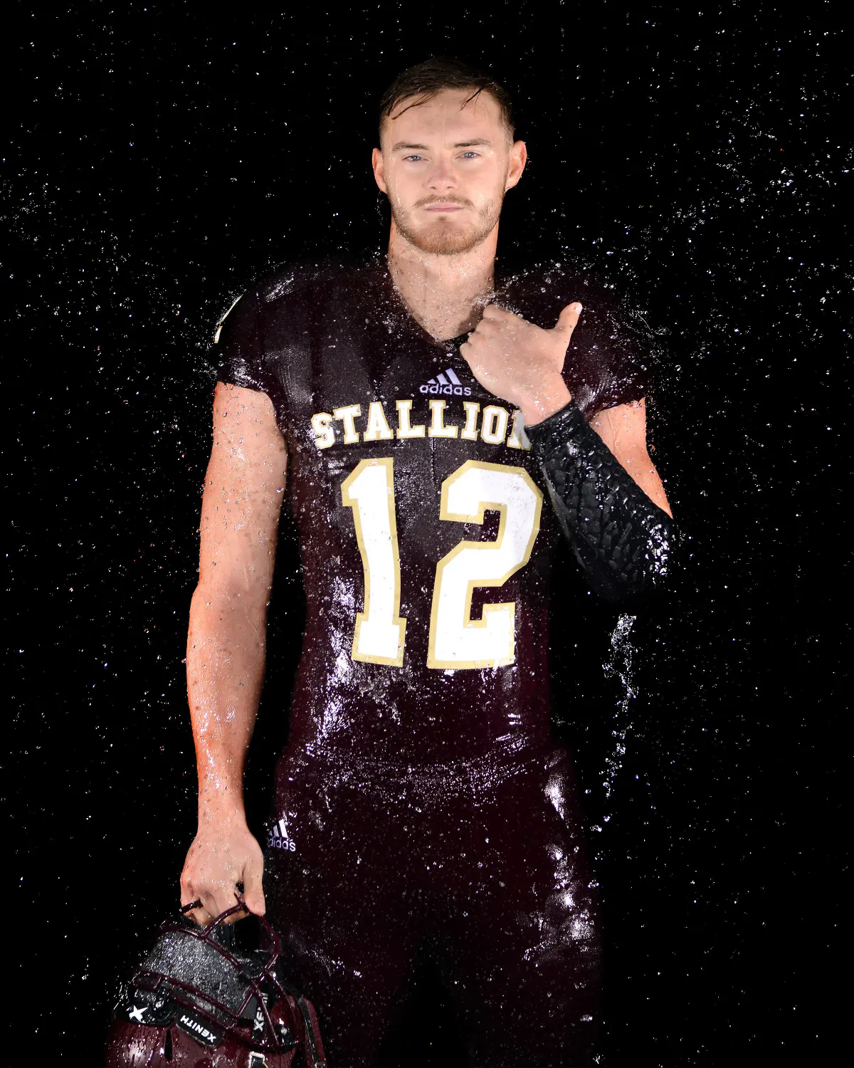 Young man in a drenched maroon 'STALLIONS 12' football jersey, hand on chest, holding a helmet at his side