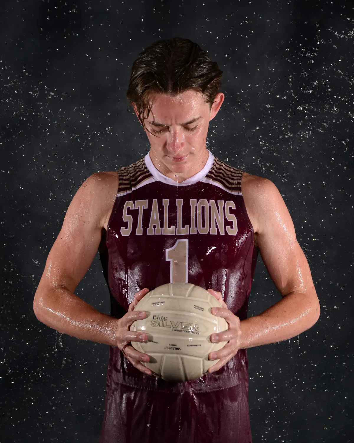 Young man in a wet maroon 'STALLIONS 1' volleyball jersey, head bowed and eyes closed, holding a volleyball at waist