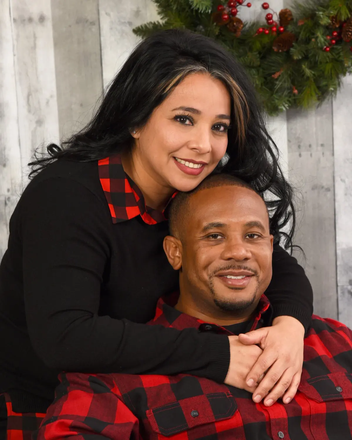 Couple holiday portrait: woman with long dark hair embracing a smiling man from behind, both in red/black buffalo-check and black