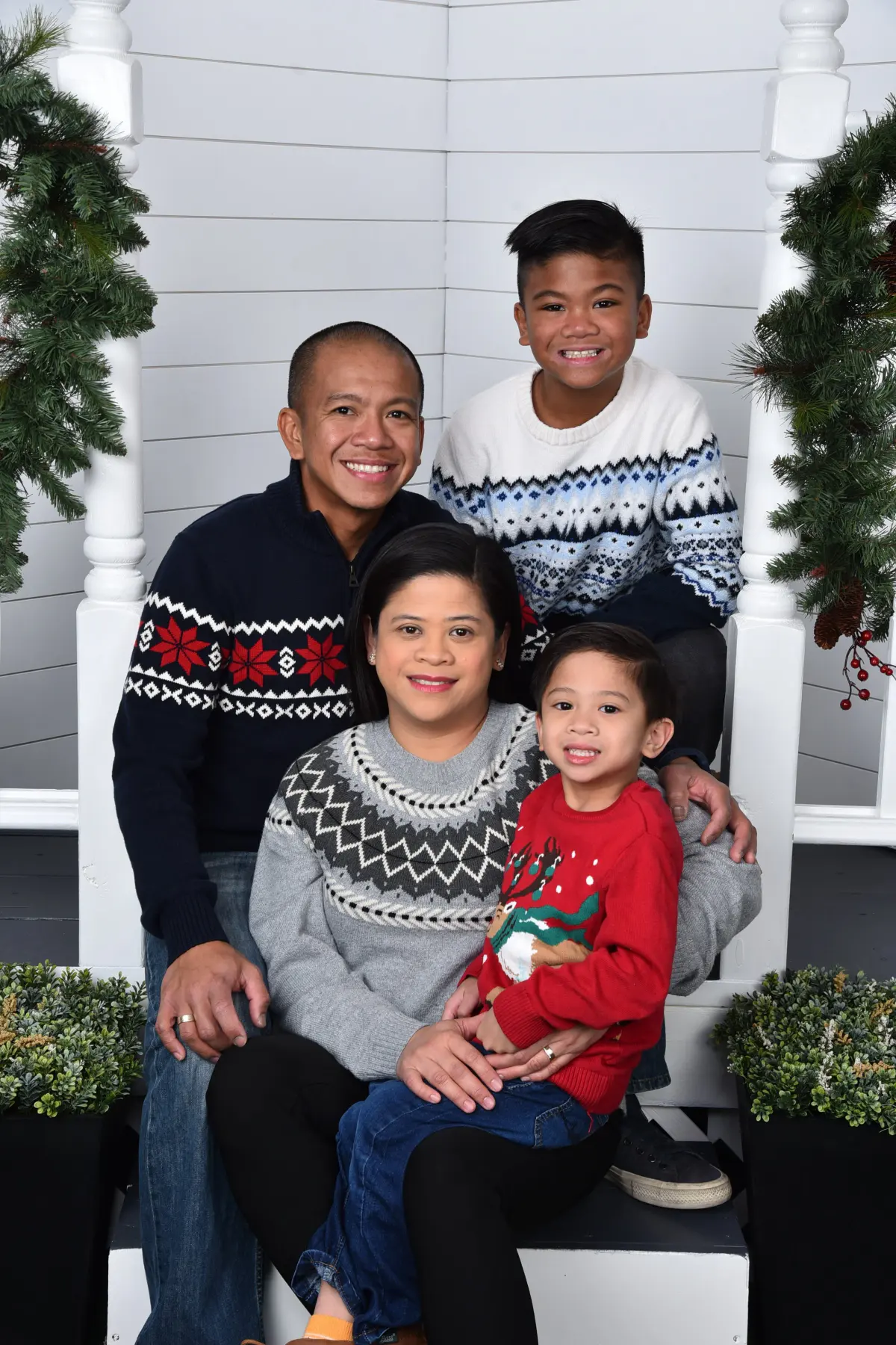 Family of four (two adults, two boys) in holiday sweaters posed on the steps of a white porch/door set with garland, wreaths and potted evergreens