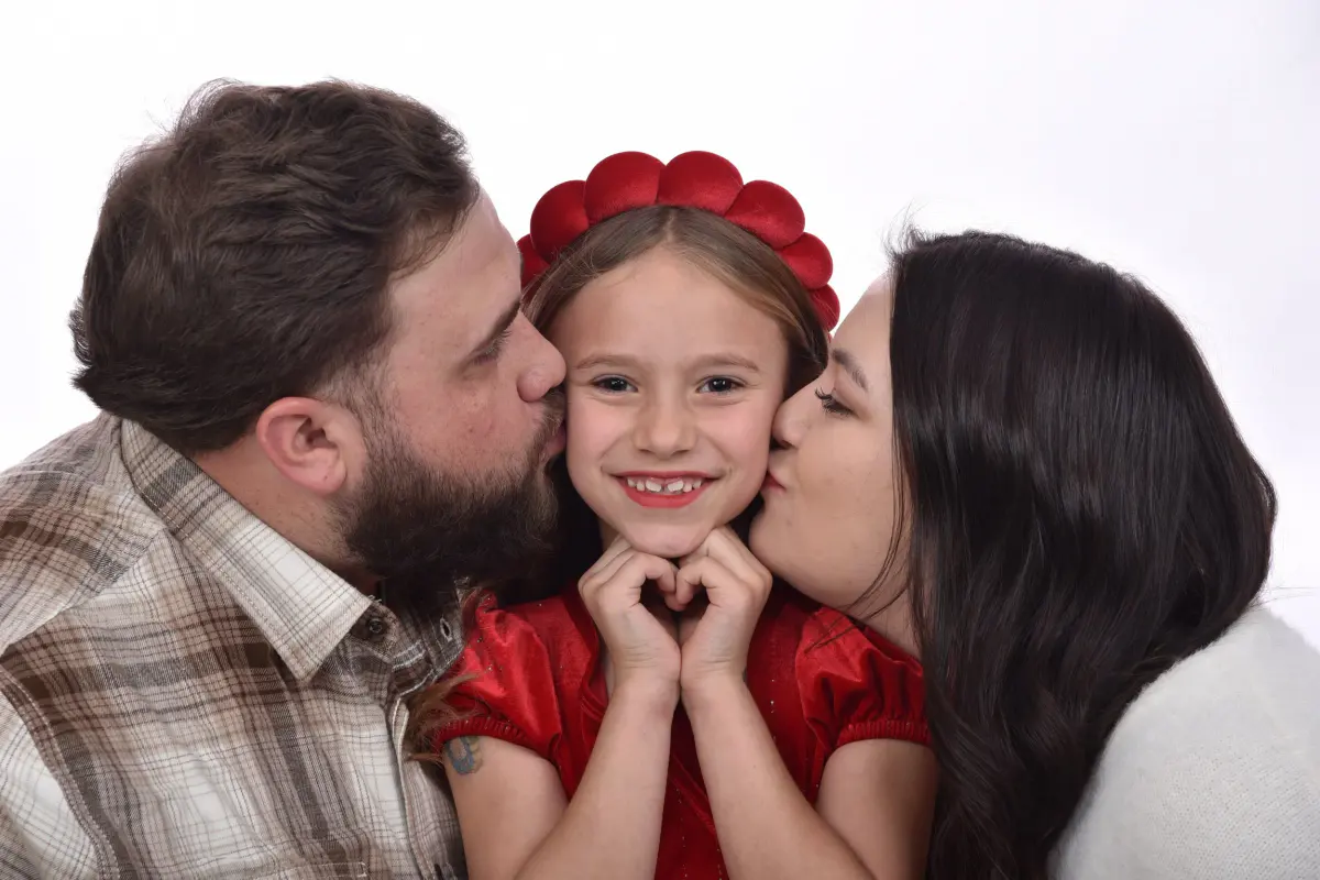 Father with beard and plaid shirt and mother with long dark hair each kissing a cheek of their young daughter