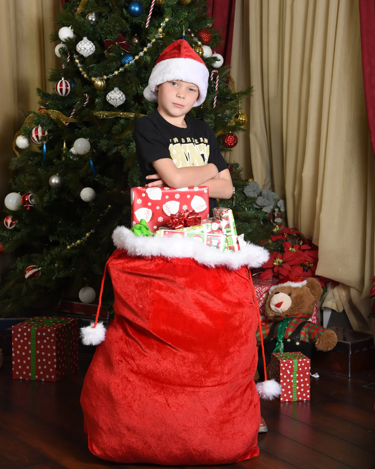 Young boy in a Santa hat and black tee, arms crossed, peeking from a large red velvet Santa sack topped with wrapped gifts; decorated Christmas tree